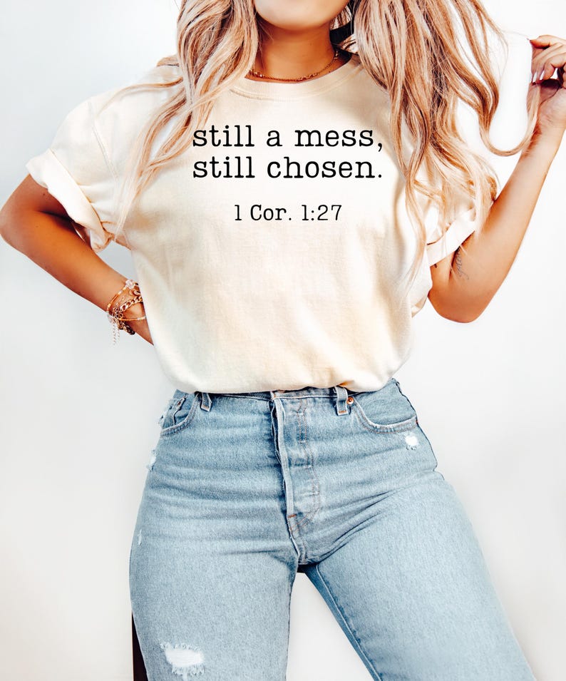 K&ouml;nnte beinhalten: Cremefarbenes T-Shirt mit dem Text "still a mess, still chosen." und dem Verweis "1 Cor. 1:27" in Schwarz. Das Shirt wird mit hellblauen Jeans kombiniert.