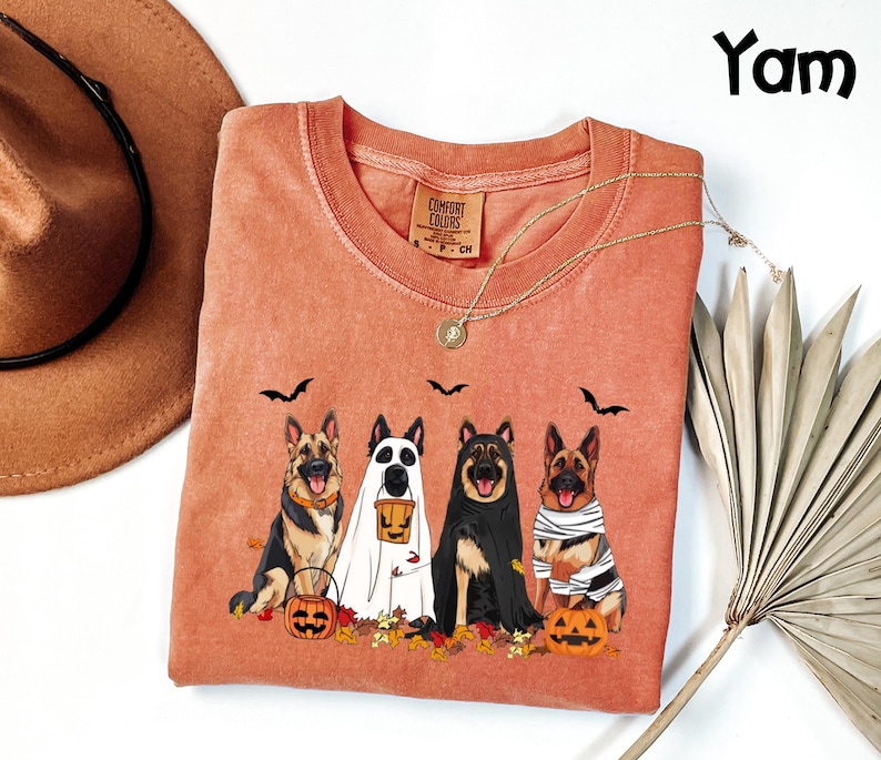 May include: A coral-colored Comfort Colors sweatshirt featuring a Halloween-themed graphic. The design includes four German Shepherds dressed as a ghost, mummy, and other costumes, with pumpkins and bats. The word "Yam" is in the upper right corner.