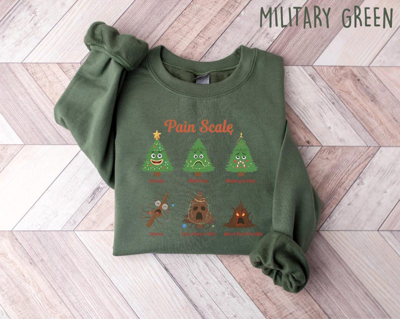 Puede incluir: Sudadera verde militar con el texto "Pain Scale" y &aacute;rboles de Navidad ilustrados que representan diferentes niveles de dolor. La sudadera es de tejido suave, con cuello redondo y mangas largas.