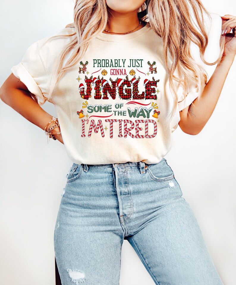 May include: Cream-colored t-shirt with the text "Probably Just Gonna Jingle Some of the Way I'm Tired" in green, red, and white. The design includes reindeer, Santa hats, bells, and candy cane stripes, perfect for the holiday season.