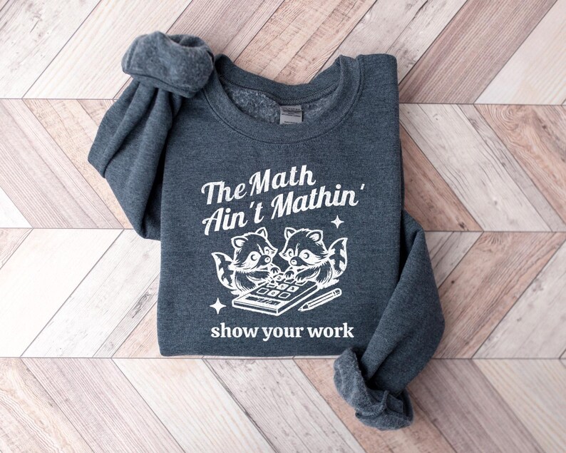 May include: A dark gray sweatshirt with a white graphic of two raccoons using a calculator. The text on the sweatshirt reads "The Math Ain't Mathin' show your work".