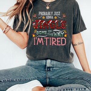 May include: Dark gray t-shirt with the text "Probably Just Gonna Jingle Some of the Way I'm Tired" in red and white. The word "Jingle" is in a plaid pattern. The shirt is worn with blue jeans and white sneakers.