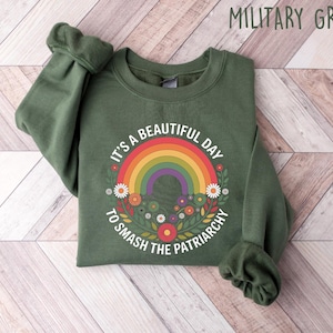 May include: Military green sweatshirt with a rainbow and floral design. The text reads "It's a beautiful day to smash the patriarchy". The sweatshirt is made of a soft material.