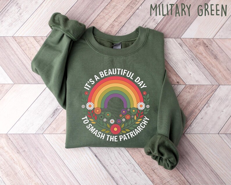 May include: Military green sweatshirt with a rainbow and floral design. The text reads "It's a beautiful day to smash the patriarchy". The sweatshirt is made of a soft material.
