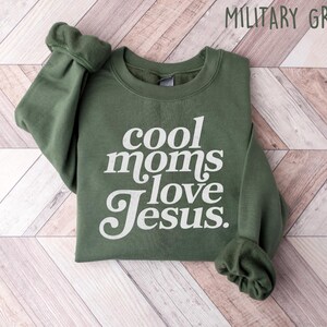 May include: Military green sweatshirt with the text "cool moms love Jesus" in white script. The sweatshirt is folded on a light wood-grain surface. The sleeves are cuffed.