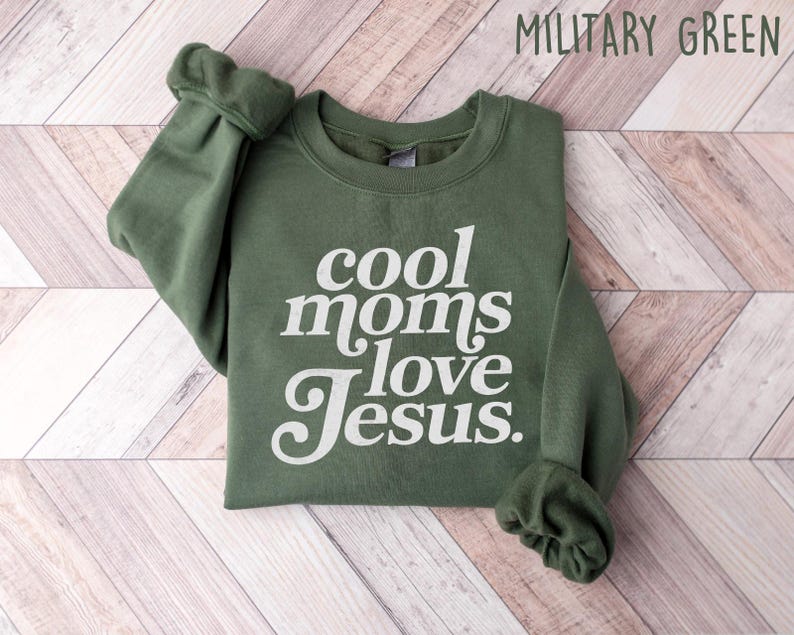 May include: Military green sweatshirt with the text "cool moms love Jesus" in white script. The sweatshirt is folded on a light wood-grain surface. The sleeves are cuffed.