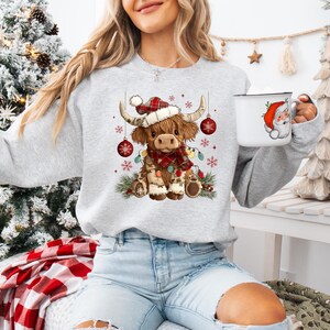 May include: A light gray sweatshirt featuring a festive Highland cow illustration. The cow wears a Santa hat, a plaid scarf, and is adorned with Christmas lights and ornaments. A white mug with a Santa Claus design is also visible.
