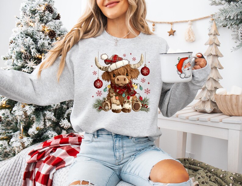 May include: A light gray sweatshirt featuring a festive Highland cow illustration. The cow wears a Santa hat, a plaid scarf, and is adorned with Christmas lights and ornaments. A white mug with a Santa Claus design is also visible.