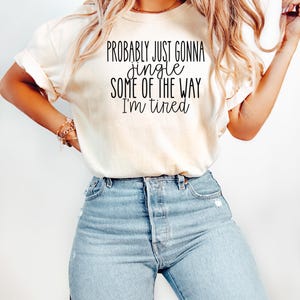 Op de afbeelding: Cr&egrave;mekleurig T-shirt met de zwarte tekst "Probably Just Gonna Jingle Some of the Way I'm Tired". Het shirt wordt gecombineerd met lichtblauwe jeans.