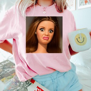 May include: Pink sweatshirt with a square image of a doll's face making a disgusted expression. A white mug with a yellow smiley face is held in the right hand. Light blue shorts and books are also visible.