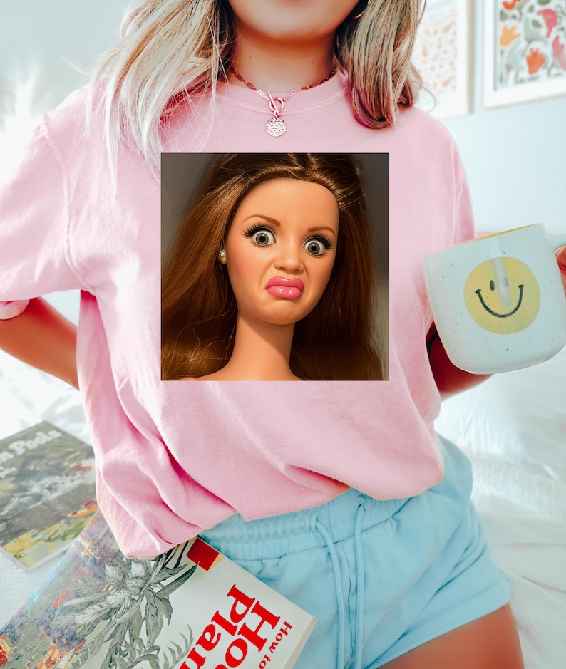 May include: Pink sweatshirt with a square image of a doll's face making a disgusted expression. A white mug with a yellow smiley face is held in the right hand. Light blue shorts and books are also visible.