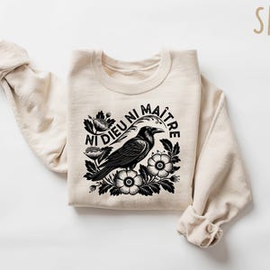 Op de afbeelding: Zandkleurige sweatshirt met een zwart-witte afbeelding van een kraai omringd door bloemen en de Franse tekst "Ni Dieu Ni Ma&icirc;tre". De top met lange mouwen is gemaakt van een zacht materiaal.