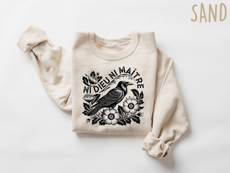 Op de afbeelding: Zandkleurige sweatshirt met een zwart-witte afbeelding van een kraai omringd door bloemen en de Franse tekst "Ni Dieu Ni Ma&icirc;tre". De top met lange mouwen is gemaakt van een zacht materiaal.