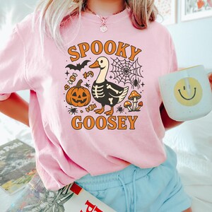 May include: Pink t-shirt with a Halloween-themed graphic. The design features a goose with a skeleton body, a jack-o'-lantern, a spider web, and the words "Spooky Goosey" in orange lettering. The person is also holding a mug with a smiley face.