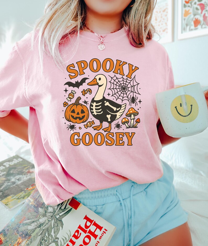 May include: Pink t-shirt with a Halloween-themed graphic. The design features a goose with a skeleton body, a jack-o'-lantern, a spider web, and the words "Spooky Goosey" in orange lettering. The person is also holding a mug with a smiley face.