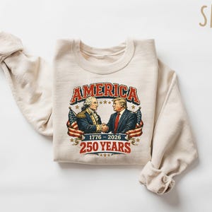 Puede incluir: Una sudadera de color arena con un gr&aacute;fico de dos hombres d&aacute;ndose la mano, con la palabra "AMERICA" encima. El gr&aacute;fico incluye el texto "1776-2026" y "250 YEARS" en un estilo vintage. Banderas americanas a ambos lados del gr&aacute;fico.