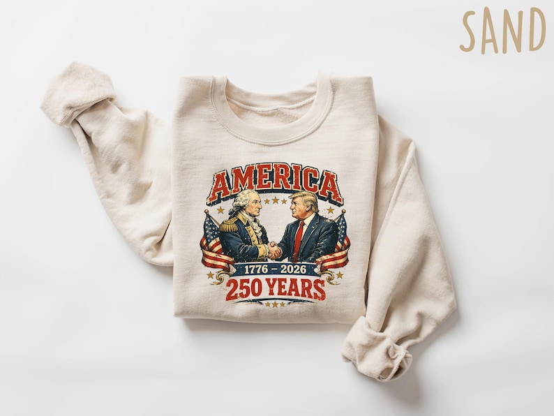 Puede incluir: Una sudadera de color arena con un gr&aacute;fico de dos hombres d&aacute;ndose la mano, con la palabra "AMERICA" encima. El gr&aacute;fico incluye el texto "1776-2026" y "250 YEARS" en un estilo vintage. Banderas americanas a ambos lados del gr&aacute;fico.