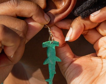 Pode incluir: Uma pessoa está colocando um brinco de martelo de tubarão verde. O brinco é feito de plástico e tem um gancho dourado.