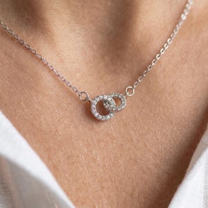 Puede incluir: Un collar de tono plateado con una cadena fina y un colgante formado por dos anillos circulares entrelazados. Ambos anillos están incrustados con pequeñas piedras claras y brillantes que captan la luz, creando un efecto resplandeciente.