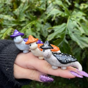 May include: Four small ceramic figurines of creatures wearing witch hats. The hats are purple, orange, black, and orange. The creatures are white with black spots and have black eyes.