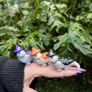 May include: Four small ceramic figurines of Halloween-themed creatures wearing witch hats. The creatures are painted in white, black, orange, and purple.
