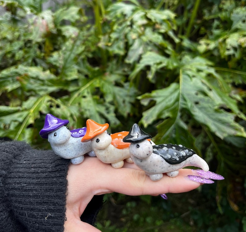 May include: Four small ceramic figurines of Halloween-themed creatures wearing witch hats. The creatures are painted in white, black, orange, and purple.