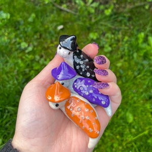 May include: Three ceramic Halloween-themed figurines, each with a different hat. The first figurine is black and white with a black witch hat. The second figurine is purple with a purple witch hat. The third figurine is orange with a black and orange witch hat.