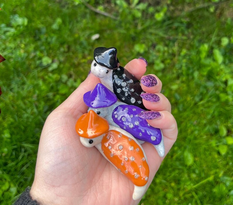 May include: Three ceramic Halloween-themed figurines, each with a different hat. The first figurine is black and white with a black witch hat. The second figurine is purple with a purple witch hat. The third figurine is orange with a black and orange witch hat.