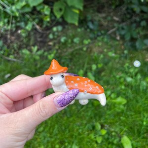 May include: A small, white ceramic figurine of a mushroom with an orange cap and white spots. The mushroom is wearing a small, orange hat.