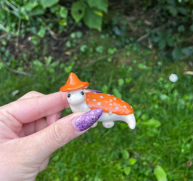 May include: A small, white ceramic figurine of a mushroom with an orange cap and white spots. The mushroom is wearing a small, orange hat.