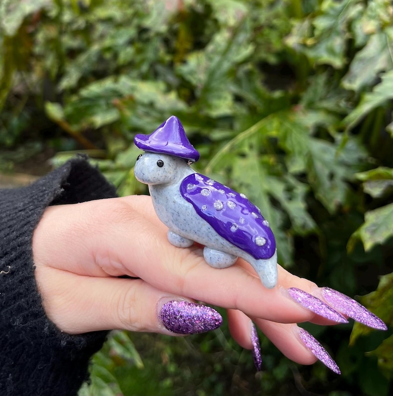 May include: A small ceramic figurine of a creature wearing a purple hat with white spots. The creature has a gray body with purple spots and a white face.
