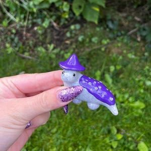 May include: A small ceramic turtle figurine wearing a purple hat with white polka dots. The turtle is painted a light gray with purple accents.