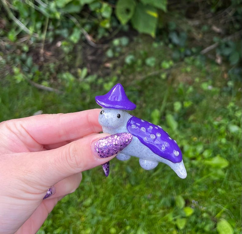 May include: A small ceramic turtle figurine wearing a purple hat with white polka dots. The turtle is painted a light gray with purple accents.