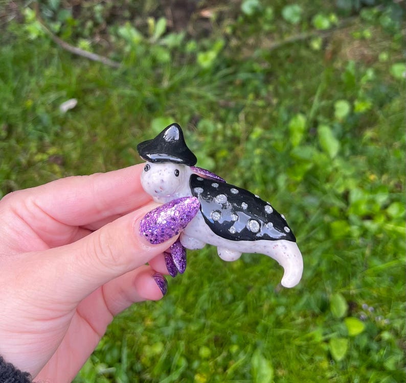 May include: A small ceramic figurine of a creature wearing a black witch hat with white polka dots. The creature is white with black spots and has a black and white striped tail.