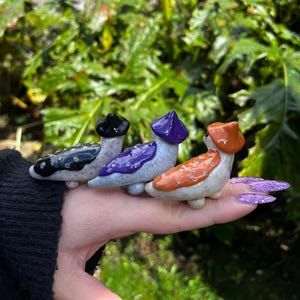 May include: Three ceramic figurines of slugs wearing different colored hats. The slugs are white with black speckles and are wearing a black, purple, and orange hat.