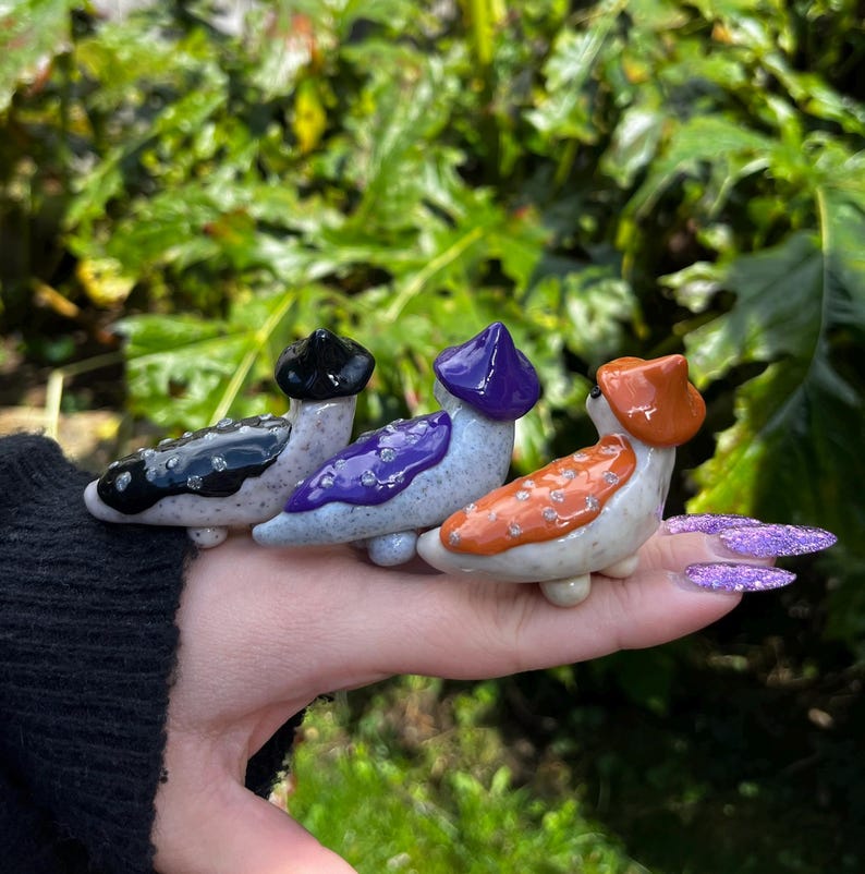 May include: Three ceramic figurines of slugs wearing different colored hats. The slugs are white with black speckles and are wearing a black, purple, and orange hat.