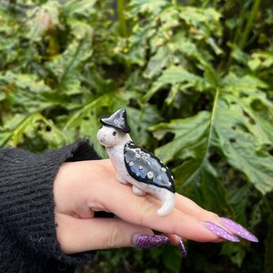 May include: A small ceramic figurine of a creature wearing a black witch hat. The creature is white with black spots and has a long tail.