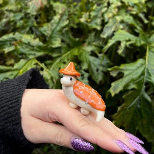 May include: A small ceramic figurine of a mushroom wearing a red and orange hat. The mushroom is white with orange spots and is standing on a person's hand.