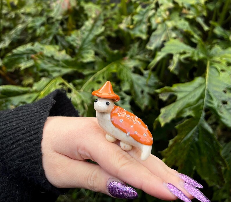 May include: A small ceramic figurine of a mushroom wearing a red and orange hat. The mushroom is white with orange spots and is standing on a person's hand.