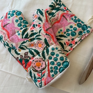 May include: A pair of oven mitts and a matching pot holder with a whimsical floral and unicorn pattern. The design features pink unicorns, coral flowers, and teal accents on an off-white background. The mitts and pot holder are quilted.