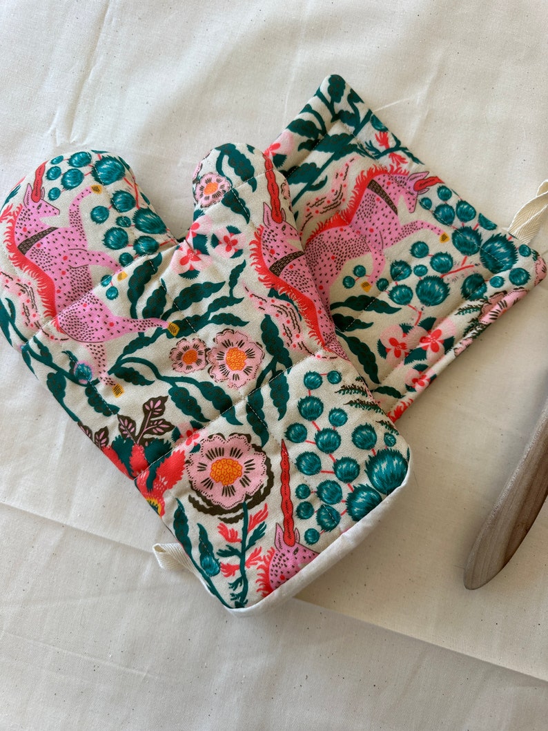 May include: A pair of oven mitts and a matching pot holder with a whimsical floral and unicorn pattern. The design features pink unicorns, coral flowers, and teal accents on an off-white background. The mitts and pot holder are quilted.