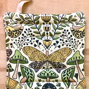 Puede incluir: Un agarrador de olla verde, amarillo y marr&oacute;n con un patr&oacute;n de mariposa y hongo. El agarrador de olla tiene un lazo para colgar.