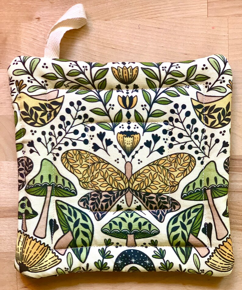 Puede incluir: Un agarrador de olla verde, amarillo y marr&oacute;n con un patr&oacute;n de mariposa y hongo. El agarrador de olla tiene un lazo para colgar.