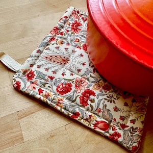May include: A floral patterned oven mitt with a loop, featuring red, yellow, and gray flowers on a cream background. A red pot rests on the mitt. The mitt is on a wooden surface.
