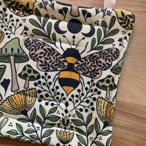 Puede incluir: Un agarrador de olla beige con un patr&oacute;n de abeja, setas, flores y hojas. El agarrador de olla tiene un lazo de color crema para colgar.