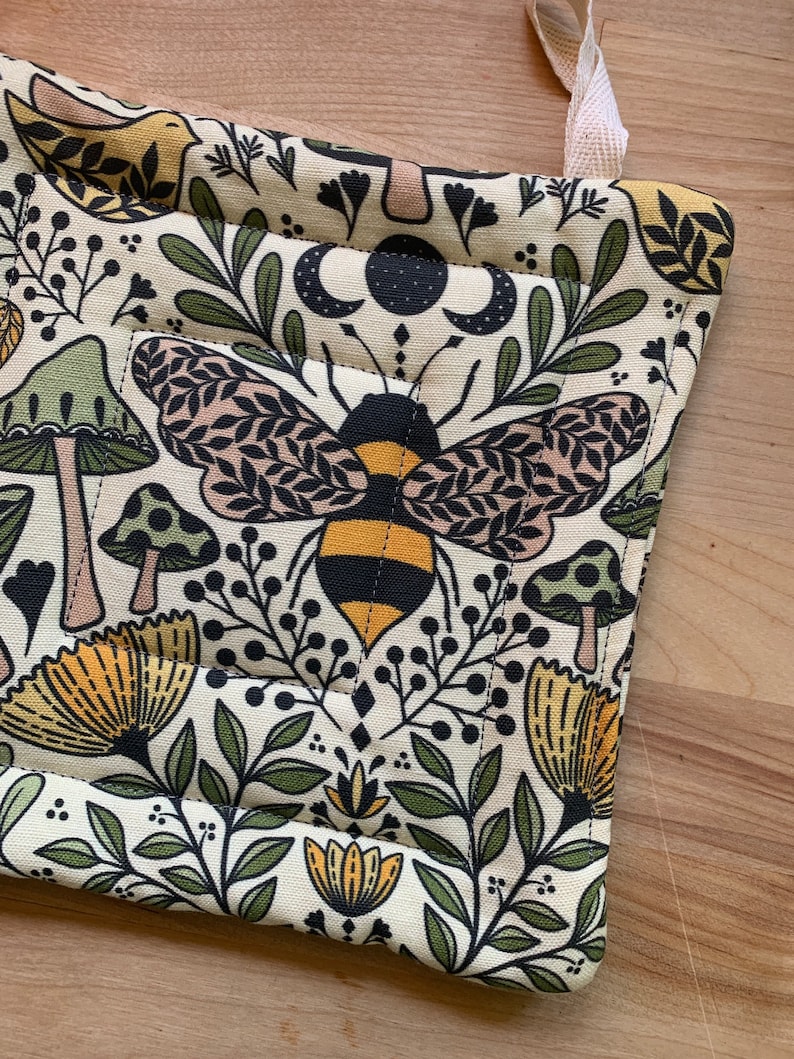 Puede incluir: Un agarrador de olla beige con un patr&oacute;n de abeja, setas, flores y hojas. El agarrador de olla tiene un lazo de color crema para colgar.