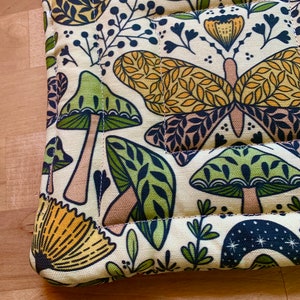 Puede incluir: Un posavasos de tela de color crema con un patr&oacute;n repetido de setas verdes, mariposas amarillas y hojas verdes. La tela est&aacute; acolchada y tiene un borde cosido.