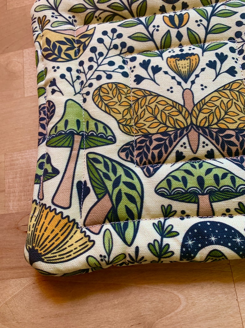 Puede incluir: Un posavasos de tela de color crema con un patr&oacute;n repetido de setas verdes, mariposas amarillas y hojas verdes. La tela est&aacute; acolchada y tiene un borde cosido.