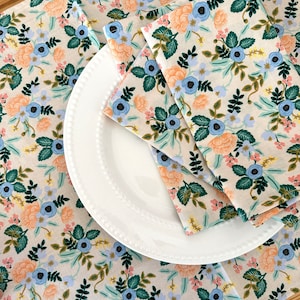 May include: A white ceramic plate with a beaded edge is layered on top of two floral patterned fabric napkins. The napkins have a light beige background with a repeating pattern of blue, pink, and green flowers.