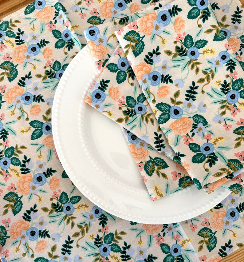 May include: A white ceramic plate with a beaded edge is layered on top of two floral patterned fabric napkins. The napkins have a light beige background with a repeating pattern of blue, pink, and green flowers.